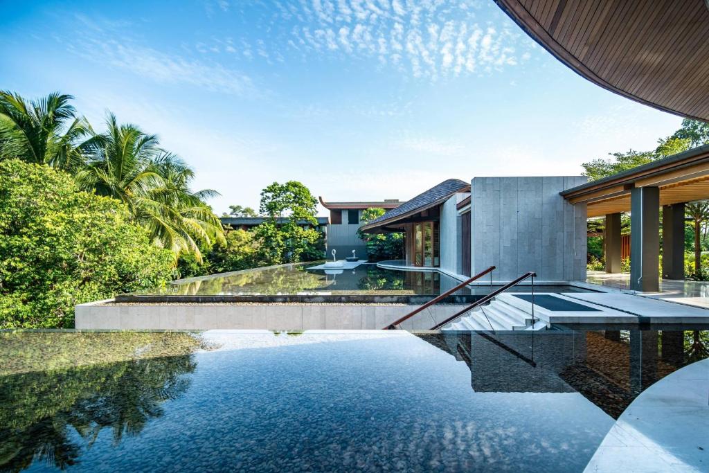 Mūsdienīga villa ar stikla baseinu un apkārt esošiem tropiskiem augiem, kas rada mierīgu un relaksējošu atmosfēru. Pilsētas ainava un skaidra debesis piešķir attēlam pievilcību.