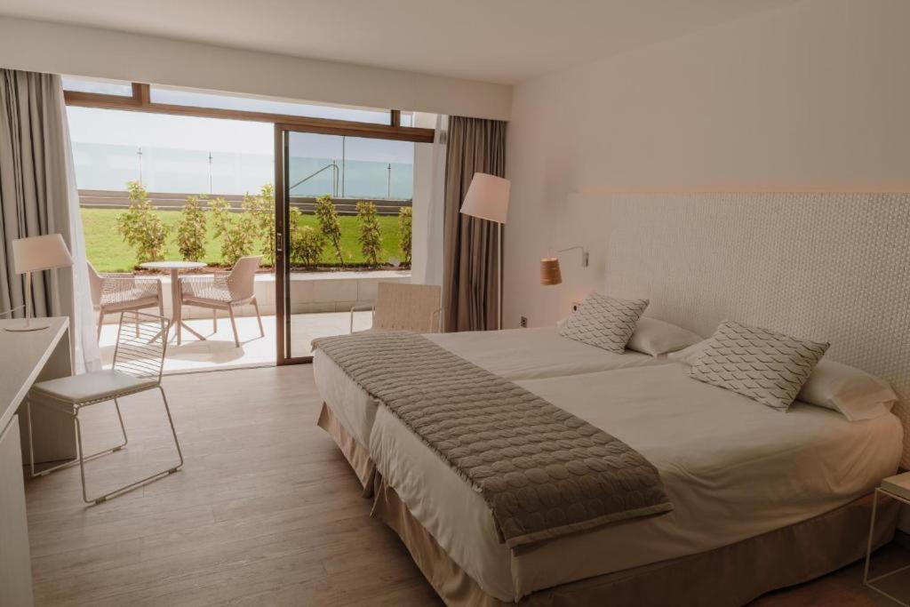 Mūsdienīgs viesnīcas numurs ar divām gultām, koka grīdu un lielu logu, kas ved uz terasi ar āra mēbelēm un zaļo zālienu.