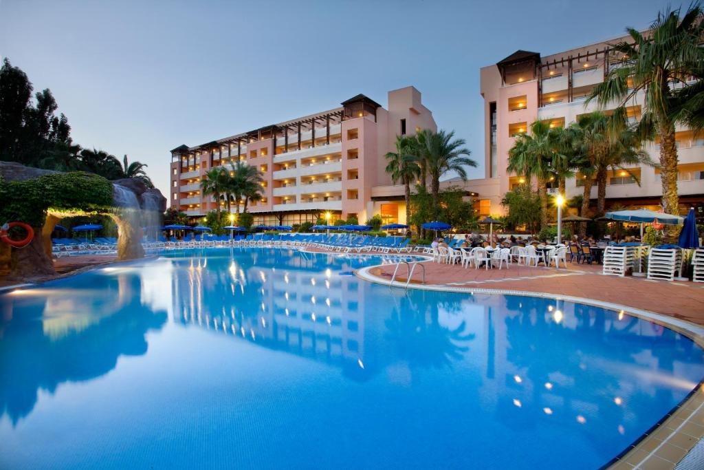 Viesnīcas baseins ar palmām un sauļošanās krēsliem, apkārt esošās ēkas un dārzi radīja relaksējošu atmosfēru.