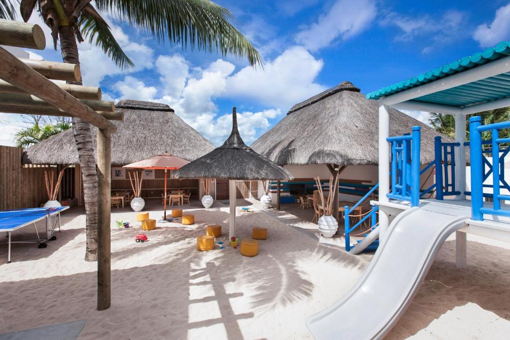 Bērnu rotaļu laukums pludmalē ar smiltīm, zilo slaidu un salmu jumtu mājiņām, kas piedāvā ērtu atpūtu un jautrību. Ideāla vieta bērnu izklaidei siltā saulē.