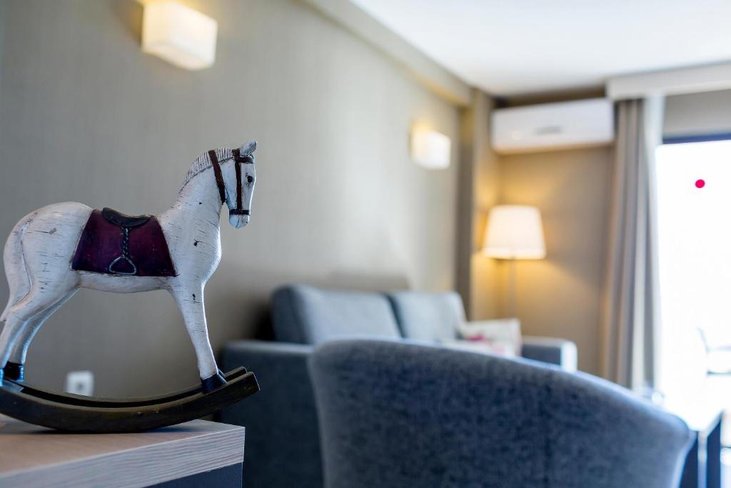 Koka šūpuļzirgs ar sarkanu sēdekli, novietots interjera telpā, ar dīvānu un gaismas avotu fona.