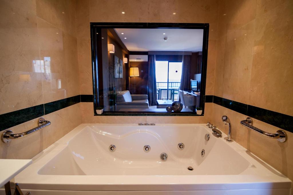 Mūsdienīga vanna ar burbuļiem, kas atrodas viesnīcas numurā, ar skatu uz viesistabu un balkonu. Elegants interjers ar neitrālām krāsām un mūsdienīgu apgaismojumu.