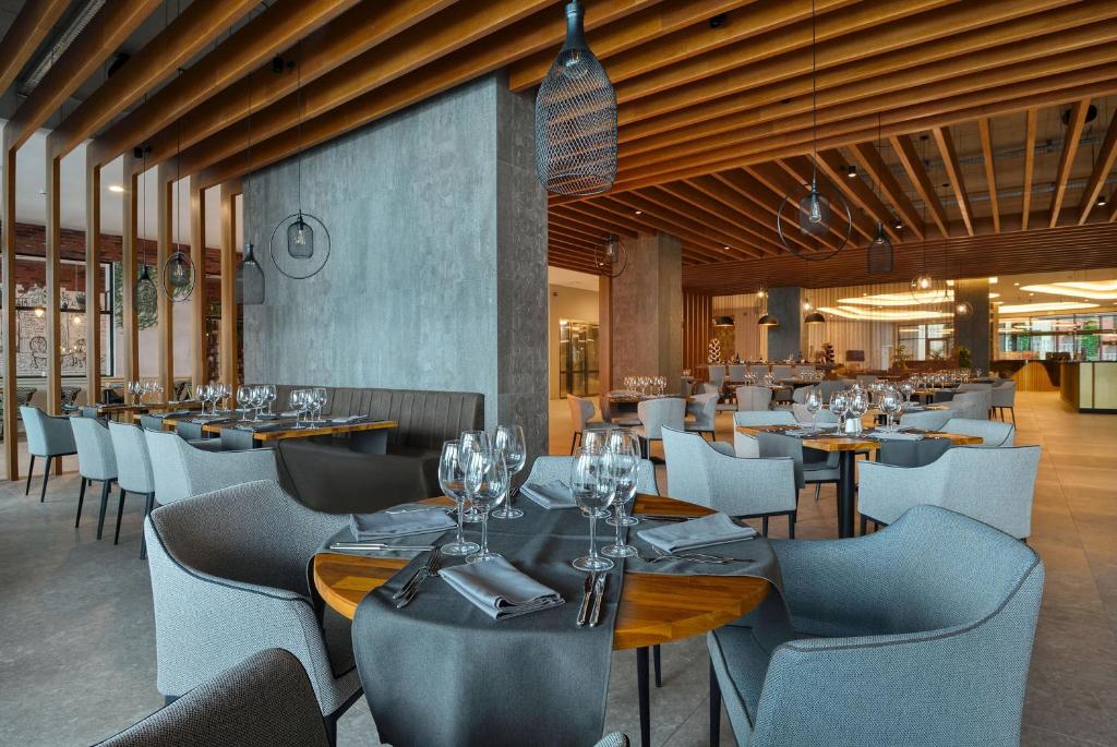 Mūsdienīga restorāna interjers ar elegantiem galdiem un krēsliem, kas izveidots ar koka un betona elementiem. Telpa ir gaiša un plaša, radot patīkamu atmosfēru ēšanai.
