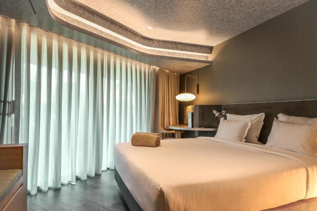 Mūsdienīgs guļamistabas interjers ar plašām logu aizkariem un ērtu gultu, kas nodrošina relaksējošu atmosfēru. Gaismas un krāsu kombinācija rada mierīgu un stilīgu vidi.