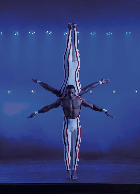 Divi akrobāti izpilda sarežģītu līdzsvara triku uz skatuves, tērpti baltos kostīmos ar sarkanām svītrām. Fons ir tumšs ar gaismas efektiem, radot dramatisku atmosfēru.