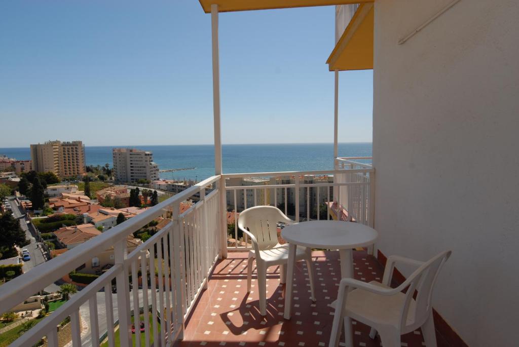 Skatījums no balkona uz jūru, kur redzams galds un divas krēsli, apkārt esošās ēkas un zaļumi. Ideāli piemērots relaksācijai un baudīšanai.