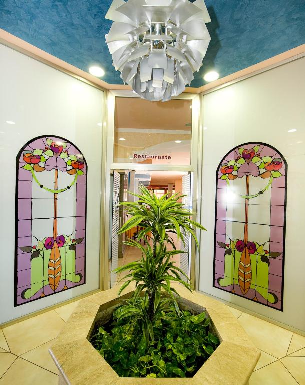 Vibrant ieeja uz restorānu ar krāsainiem vitrāžas logiem un zaļu augu centrā. Moderns apgaismojums papildina viesmīlīgo atmosfēru.