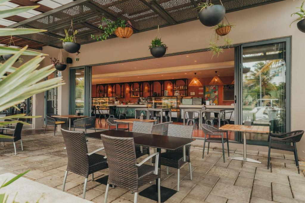 Kafejnīcas terase ar modernu dizainu, kurā redzami eleganti galdi un krēsli, kā arī dekoratīvi augi. Iekšpusē atrodas kafejnīcas bārs un kafijas pagatavošanas zona.