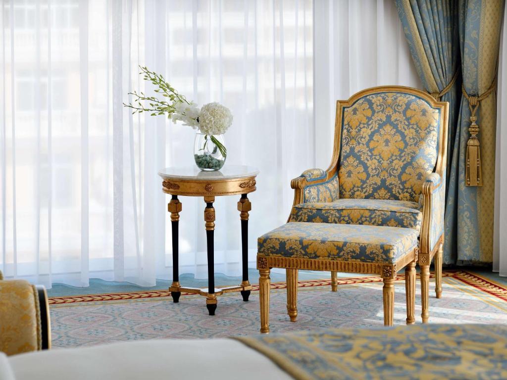 Eleganta viesistaba ar zilu un zeltītu krēslu, kas papildināts ar mazu galdiņu un ziedu kompozīciju. Gaismas aizkari rada vieglu un gaisīgu atmosfēru.