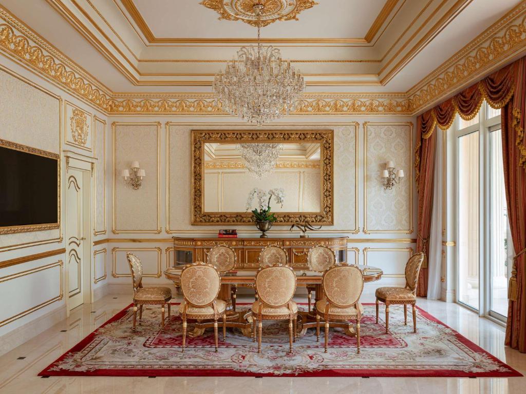 Greznas viesistabas interjers ar zelta apdari, lielu lustru un elegantu galdu ar krēsliem, kas izvietoti uz greznas paklāja. Sienas rotā izsmalcināti raksti un spogulis, radot elegantu un viesmīlīgu atmosfēru.