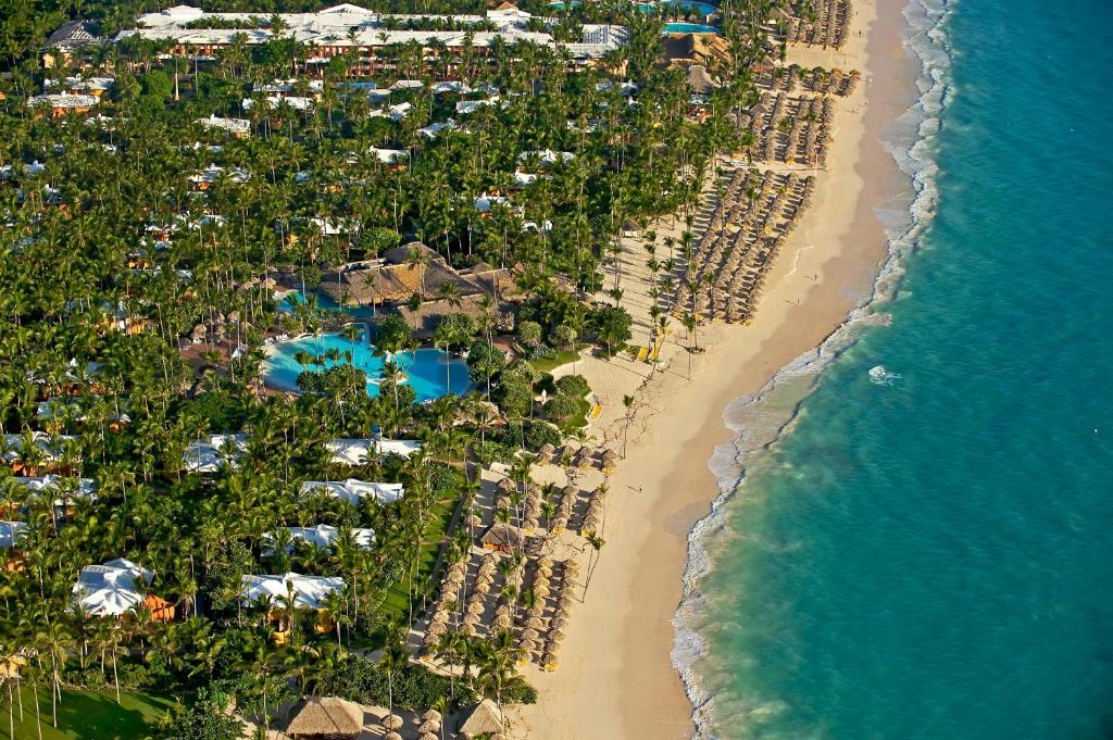 Tropiska kūrorta panorāmas skats ar zaļiem palmu kokiem, peldbaseinu un pludmali, kas stiepjas gar zilajiem ūdeņiem. Ideāla vieta atpūtai un relaksācijai.