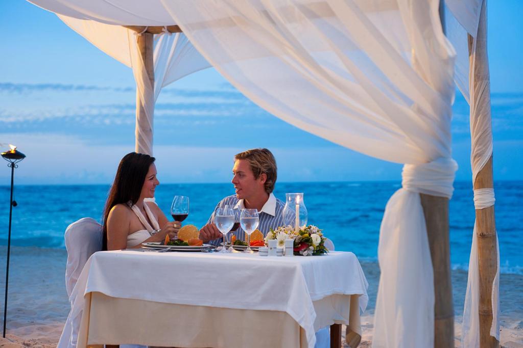 Romantiska vakariņas pludmalē zem baltiem audumiem, ar skatu uz jūru un saulrietu. Galda klāšana ar vīnu un ziediem, radot intīmu atmosfēru.