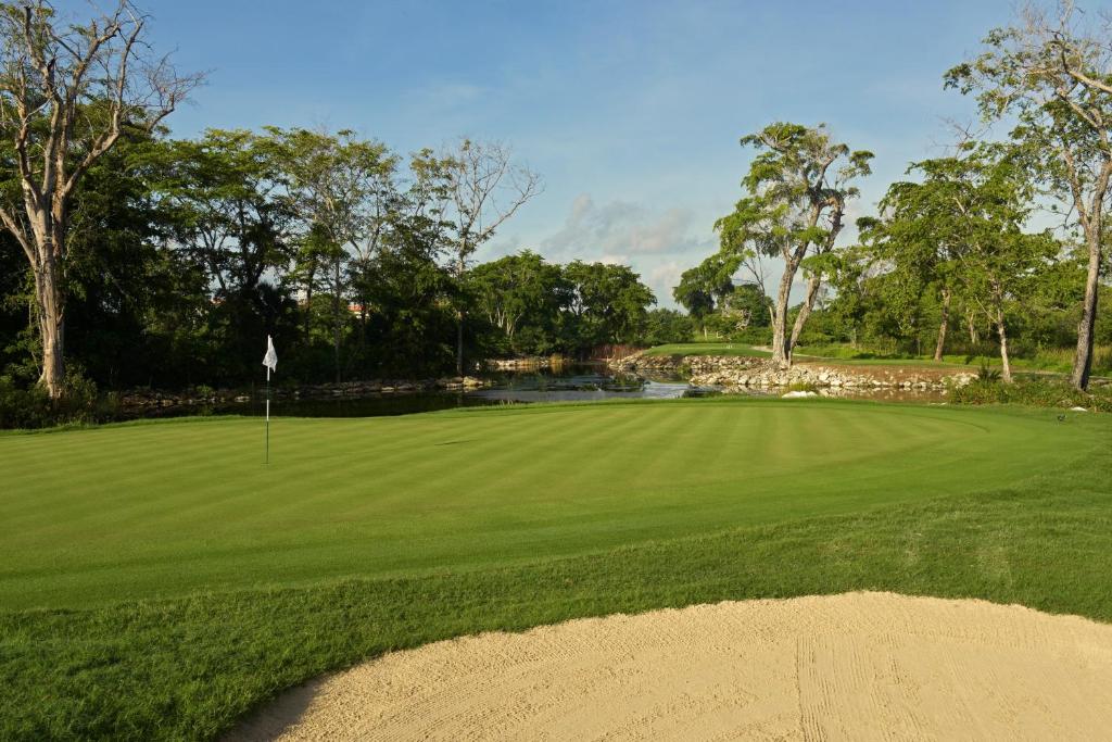 Zaļš golfa laukums ar balto golfa karogu, apņemts ar kokiem un ūdens krātuvi fona. Ideāls skats golfa spēlēšanai.