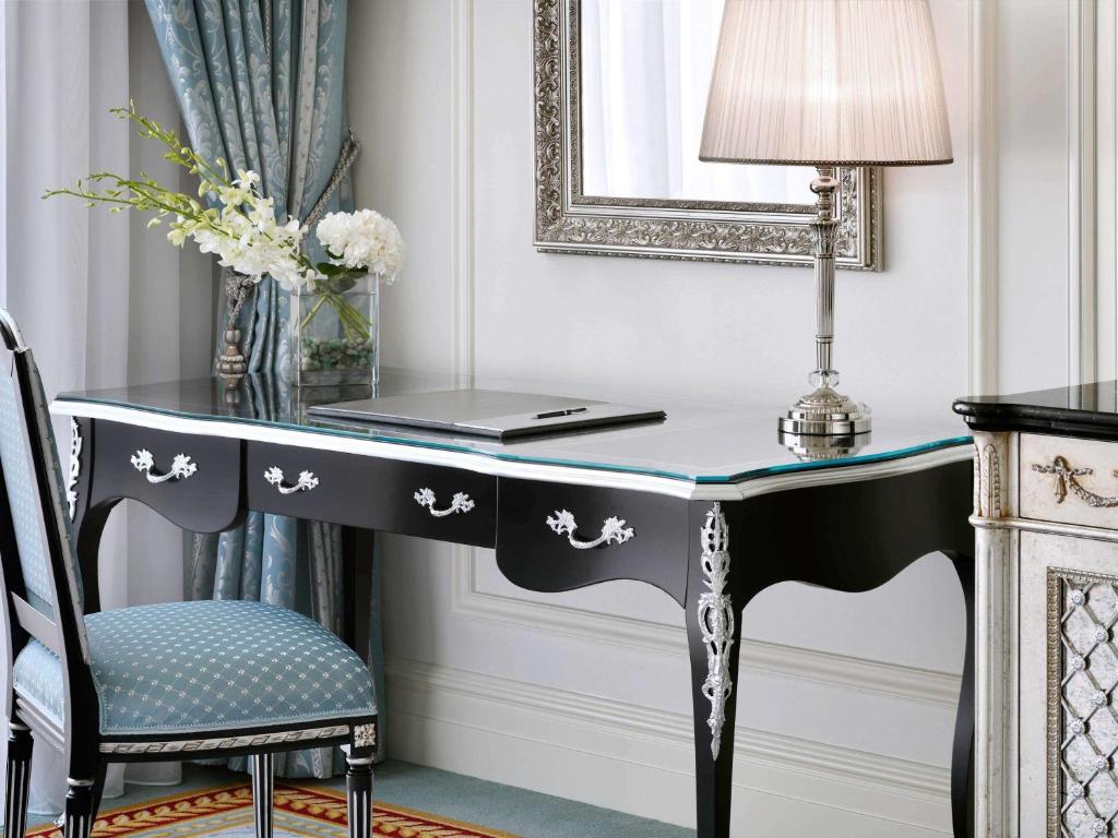 Elegants rakstāmgalds ar melnu virsmu un sudraba detaļām, blakus atrodas moderna lampa un ziedu kompozīcija. Interjerā ir redzams arī spogulis un grezni aizkari.