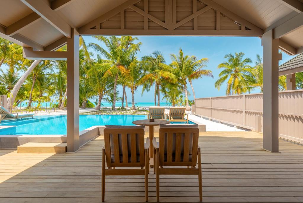 Atpūtas zona ar skatu uz baseinu un palmām, kas atrodas pie pludmales. Koka krēsli un galds aicina uz relaksāciju tropiskā vidē.