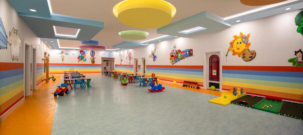 Krāsaina bērnu spēļu telpa ar zemu mēbeļu novietojumu, ko rotā spilgti zīmējumi un rotaļlietas. Telpā ir dažādi galdi un rotaļu elementi, kas veicina bērnu radošumu un izklaidi.