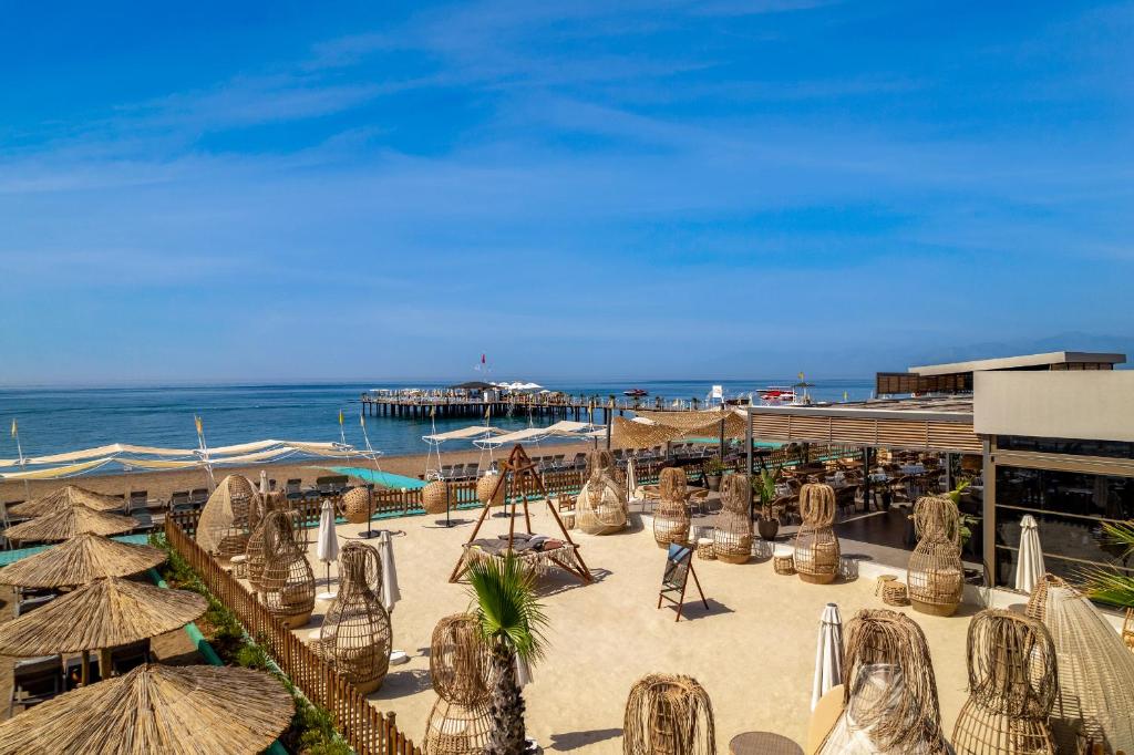 Pludmales atpūtas vieta ar smiltīm, sauļošanās krēsliem un koka konstrukcijām, kas sniedzas līdz jūrai. Fons ar skatu uz piestātni un skaidru zilo debesi.