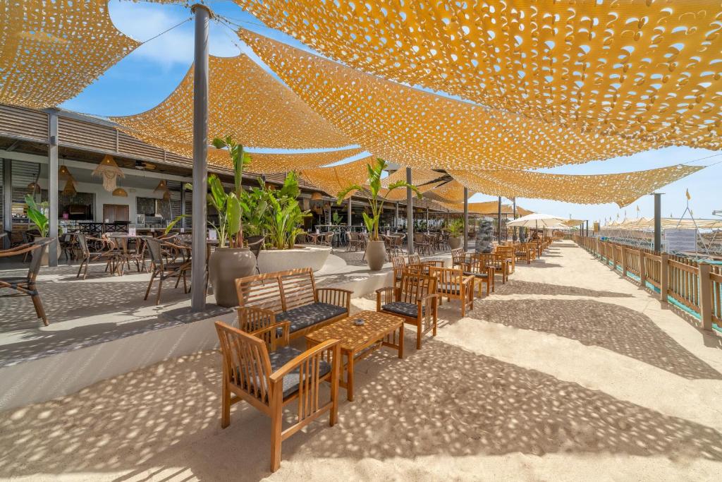 Pludmales restorāna terase ar dzelteniem auduma vairogiem, kas nodrošina ēnu, un koka mēbelēm. Apkārtne ir zaļa ar augiem, kas rada relaksējošu atmosfēru.