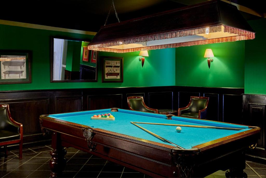Billiard room ar zaļām sienām un elegantu biljarda galdu, apgaismots ar piekārtu lampu. Galda virsma ir zila, uz tās ir biljarda bumbiņas.