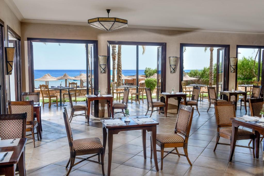 Gaisīga restorāna iekārtojums ar skatu uz jūru, atverot durvis uz terasi, kur redzamas palmas un sauļošanās krēsli. Elegantās koka mēbeles un akcenti piešķir viesmīlīgu atmosfēru.