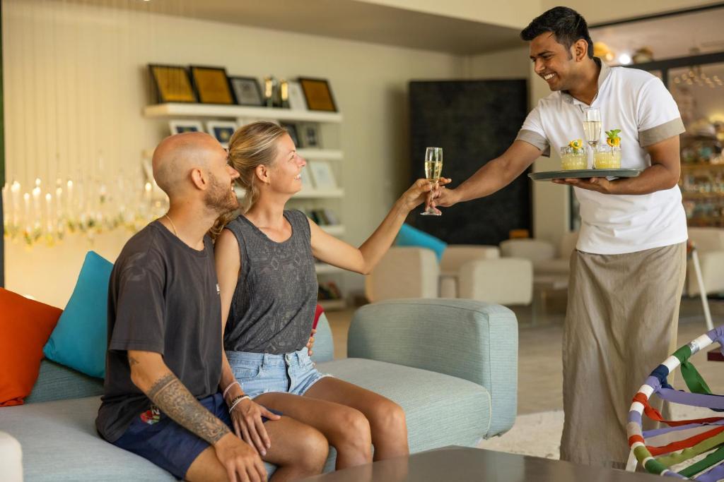 Pāris sēž viesnīcas telpā, pacelot glāzes, lai sveiktu viesmīli, kurš pasniedz dzērienus uz paplātes. Telpa ir gaiša un moderna, ar komfortablām mēbelēm un krāšņām dekora detaļām.