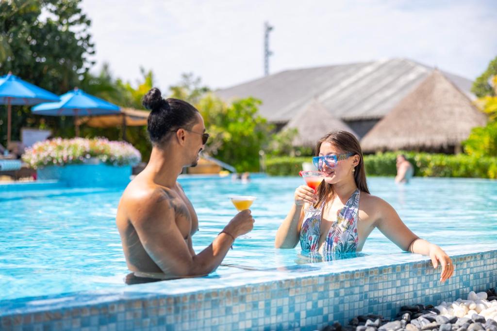 Divi cilvēki atpūšas pie baseina, baudot kokteiļus, saulainā un relaksējošā vidē. Apkārt ir ziedi un palmām, radot tropisku atmosfēru.