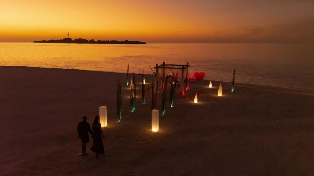 Romantiska skats uz pludmali ar pāri, kas staigā saulrieta gaismā, apgādāti ar gaismas elementiem un dekorācijām, kas rada siltu un mīlestības atmosfēru.