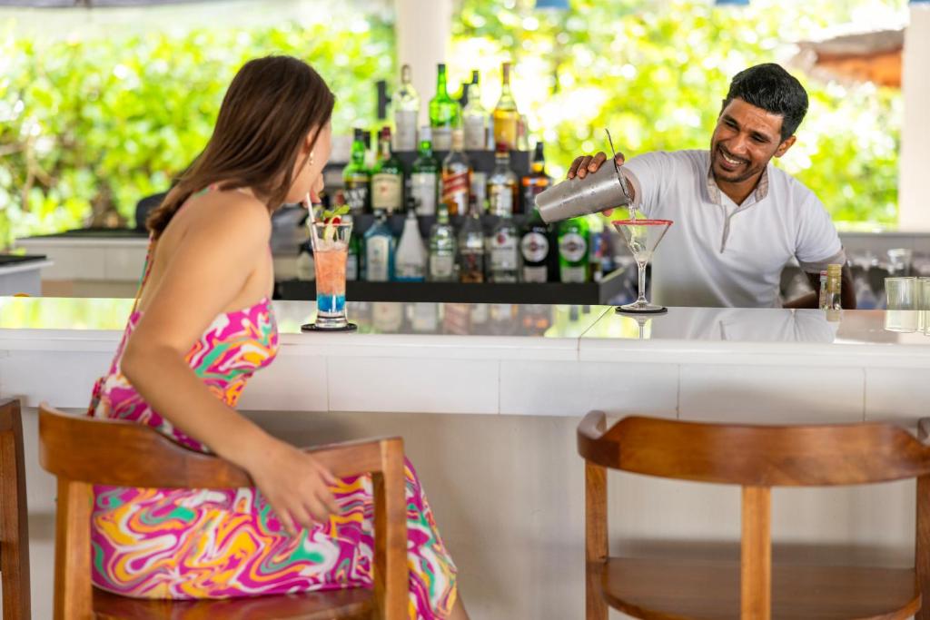 Bāra ainava, kur bārmenis pasniedz kokteili sievietei, kas sēž pie letes. Fons ar dzērienu plauktu un zaļu dabu.