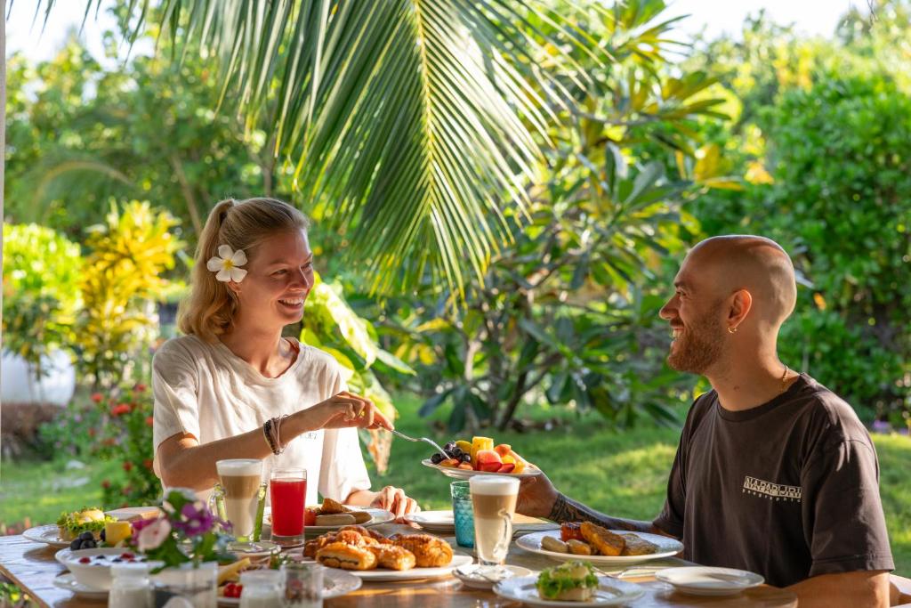 Pāris bauda brokastis tropiskā vidē, ar dažādiem ēdieniem un dzērieniem uz galda. Apkārt ziedi un zaļa daba rada patīkamu atmosfēru.