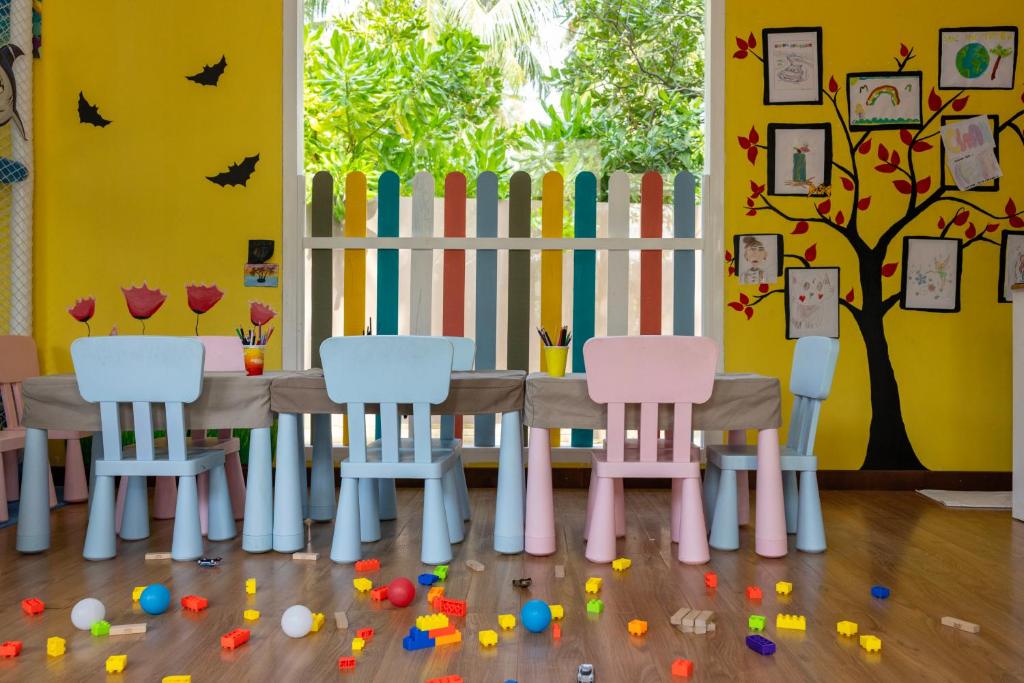 Krāsaina bērnudārza telpa ar zilu un rozā krēsliem, dekorēta ar ziedu zīmējumiem un rotaļlietām uz grīdas. Pie sienas ir krāsains koka žogs un izkārtojums ar bērnu zīmējumiem.