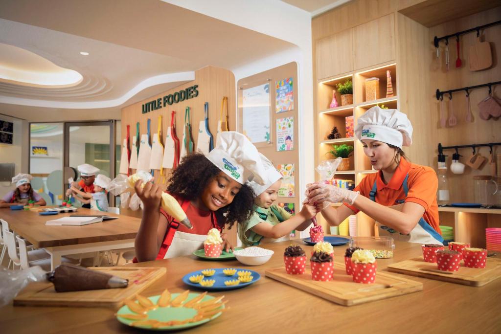 Bērni piedalās kulinārijas nodarbībā, dekorējot cepumus un saldējumus, apģērbuši šefpavāra cepures un priekšautus. Radošā virtuvē ir krāsaini trauki un dažādi gatavošanas piederumi.
