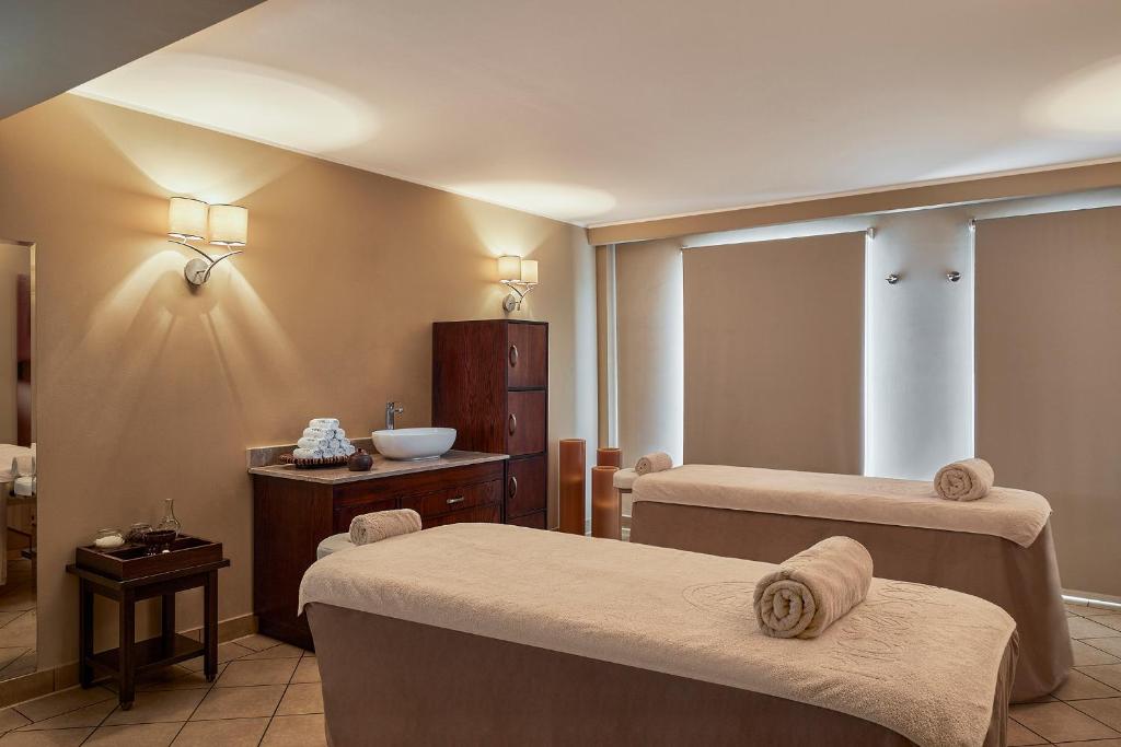 Relaksējoša spa telpa ar divām masāžas galdiem, kas rotāti ar dvieļiem, un mierīgu, siltu apgaismojumu. Fons ar koka mēbelēm un noslēgtām žalūzijām veido mierīgu atmosfēru.