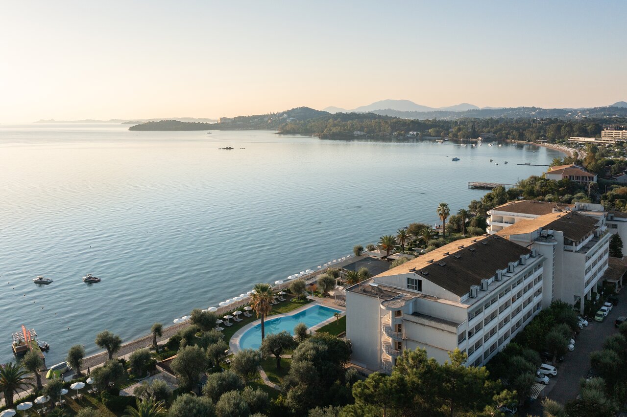 Viesnīcas skats pie jūras ar baseinu un palmām, fons ar kalniem un saulrietu. Mierīgs ūdens un laivas rada relaksējošu atmosfēru. panoramic view with beach and pools