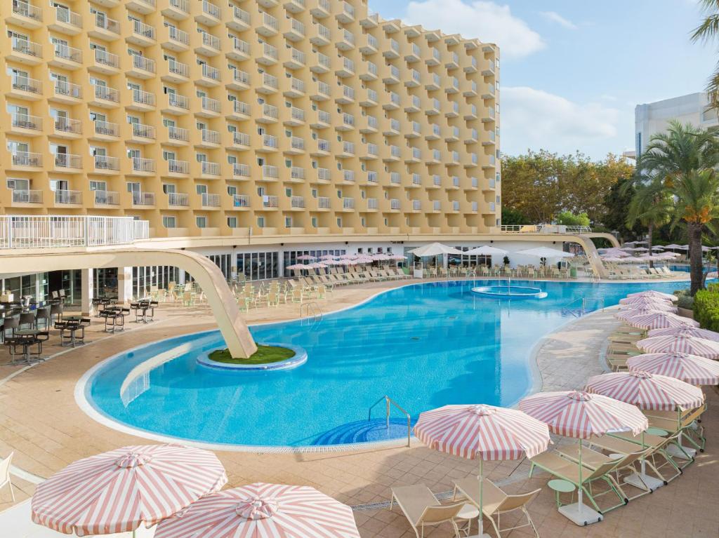 Viesnīcas baseins ar saules krēsliem un lietussargiem, apkārtne ar dzeltenu ēku un zaļām palmām. Ideāla vieta atpūtai un relaksācijai.