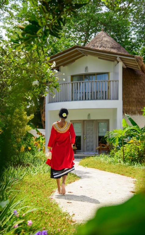 Sieviete sarkanā apģērbā pastaigā pa takti uz māju ar jumtu, ko rotā zāle, apjozta ar zaļām augiem un ziediem.