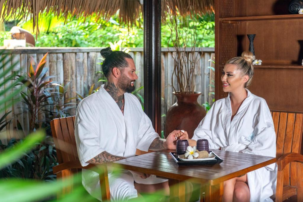 Pāris relaksējas spa vidē, ģērbušies baltos halātā, baudot mirkli kopā un turēdami viens otra rokas. Galdiņā ir izvietoti akmeņi un ziedi, radot mierīgu atmosfēru.