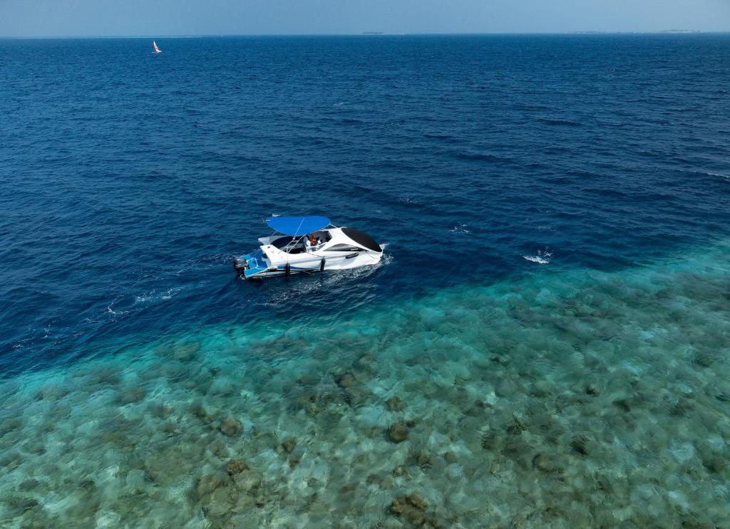 Baltā jahta peld pa skaidru, zilā jūrā, kur redzami akmeņi zem ūdens. Fons ir saulains un mierīgs, radot relaksējošu atmosfēru.