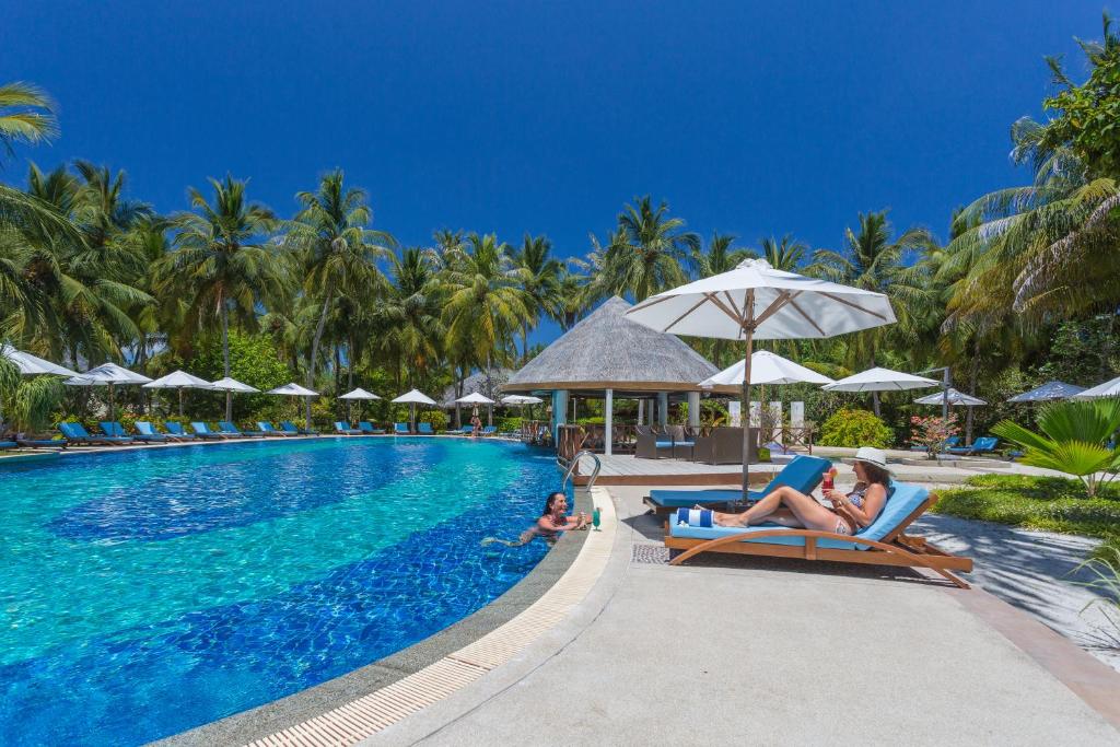 Vasaras atpūtas vieta pie baseina ar palmām un sauļošanās krēsliem, kur cilvēki relaksējas un izbauda sauli. Skaists zila ūdens baseins un mājīga ēka ar jumtu.