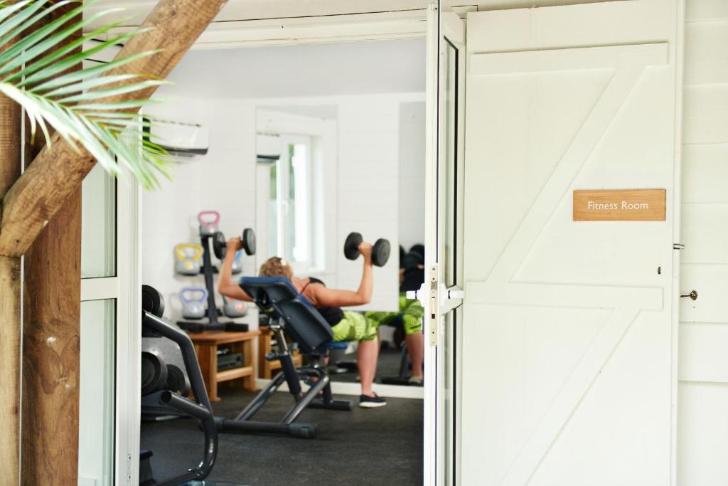 Fitnesa telpa ar aprīkojumu, kurā cilvēks trenējas ar hantelēm. Redzami dažādi vingrošanas rīki un komfortabla vide, kas veicina fizisko aktivitāti.