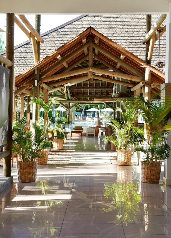 Skaista un moderna brīvdienu kūrorta ieeja ar koka konstrukcijām un zaļiem augiem, kas rada relaksējošu atmosfēru. Gaisma atspoguļojas spīdīgajos grīdas flīzēs, radot patīkamu noskaņu.