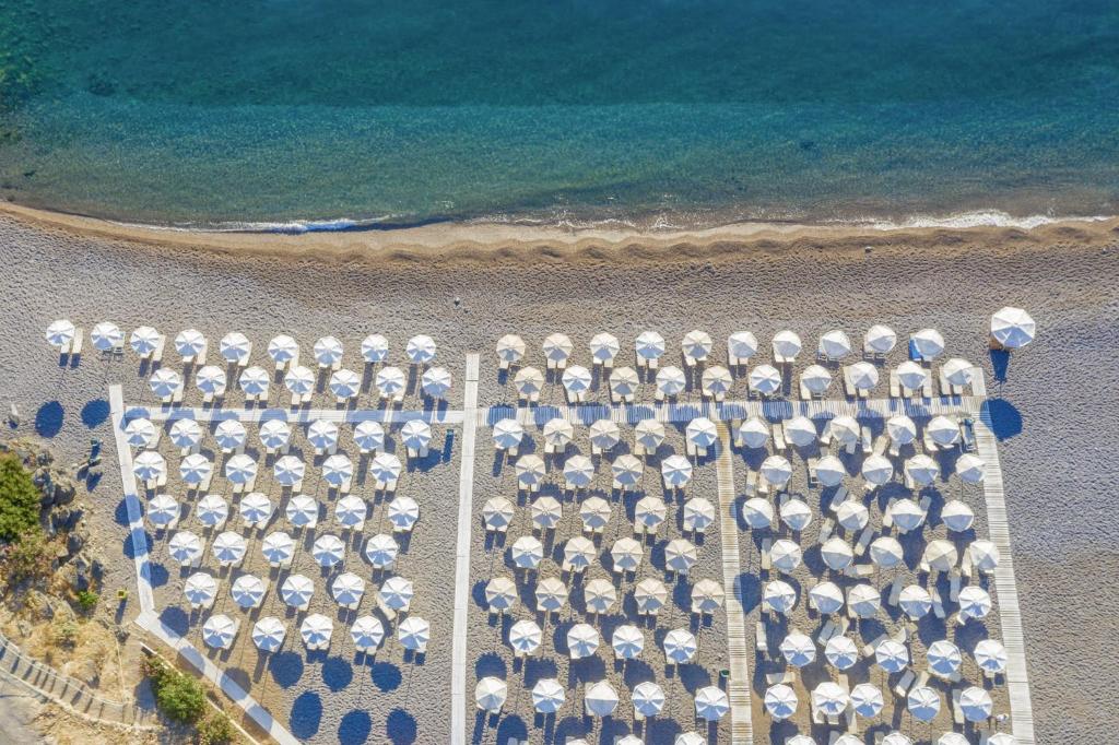 Pludmales skats no augšas, kur redzami balti saules sargi, kas sakārtoti uz akmeņainas pludmales pie zilām jūras ūdenim.