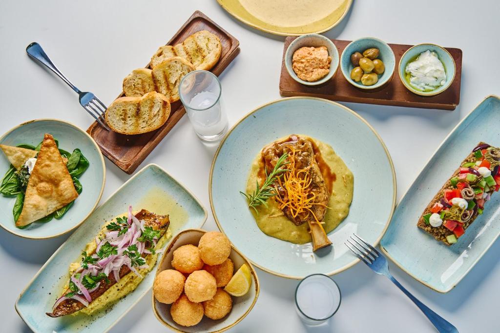 Dažādu ēdienu izkārtojums uz galda, tostarp gaļas, zivju ēdieni, pildītās maizītes un garnīri. Blakus ir dzērieni un mērces.