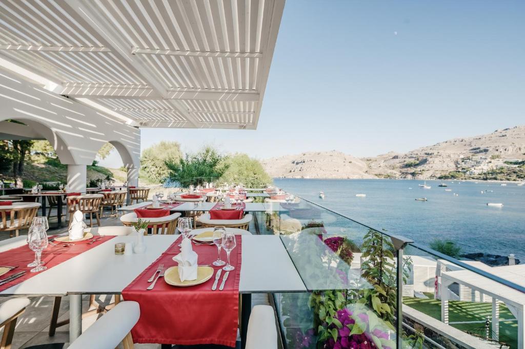 Elegantā restorāna terase ar skatu uz jūru, kur galds klāts ar sarkanām galdautām un glāzēm. Apkārt redzami kuģīši un ainaviskas kalnu ainavas.