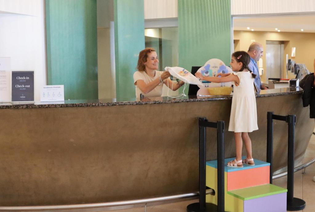 Bērns, stāvot uz krāsainām pakāpieniem, saņem dāvanu no viesnīcas reģistratūras darbinieces. Interjers ir mūsdienīgs un draudzīgs, radot patīkamu atmosfēru.