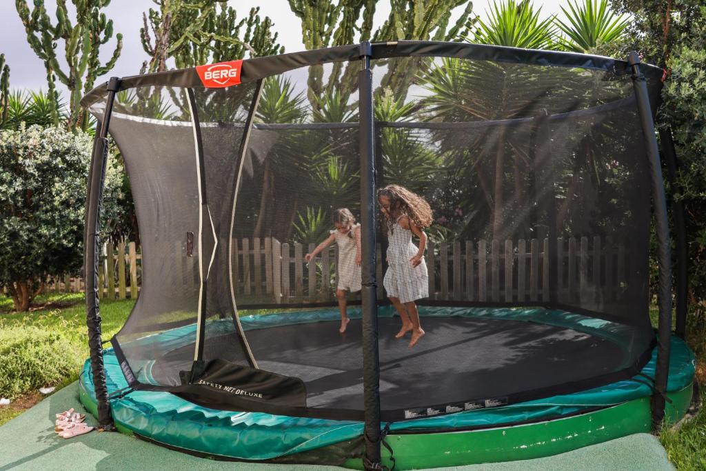 Divas meitenes priecīgi lēkā uz trenažiera ar drošības tīklu, apņemti ar zaļām palmām un žogu. Ideāla vieta bērnu izklaidei un aktīvai atpūtai.