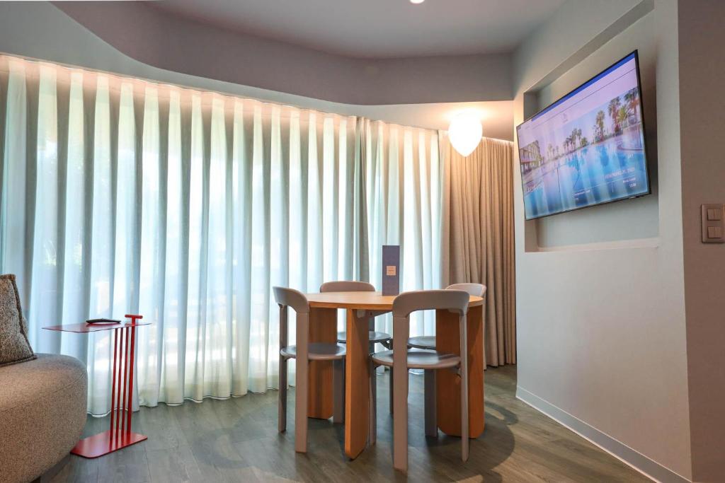 Moderns viesnīcas numuriņš ar ērtu galdu un krēsliem, lielu televizoru un gaisīgu logu ar aizkariem. Interjers ir elegants un mājīgs, radot patīkamu atmosfēru.