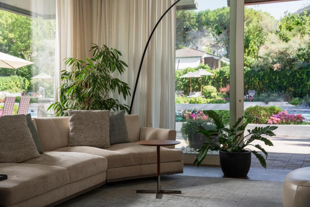 Mūsdienīgs dzīvojamās telpas interjers ar dīvānu, dekoratīviem spilveniem un stāvošu augu, kas piedāvā skatu uz dārzu ar baseinu un saules krēslām.