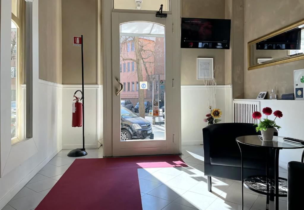 Gaišs un mūsdienīgs interjers ar sarkanu paklāju, melniem krēsliem un ziediem uz galda. Logi sniedz gaišu skatu uz ārpusi, kur redzami automobiļi un ielas aktivitāte.