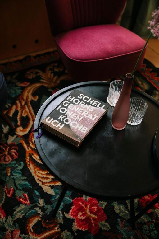 Uz melna galda atrodas grāmata ar lieliem burtiem un divas glāzes, blakus ir stilīga vāze ar ziediem. Fons veidots no krāsainas paklāja ar ziedu rakstu.