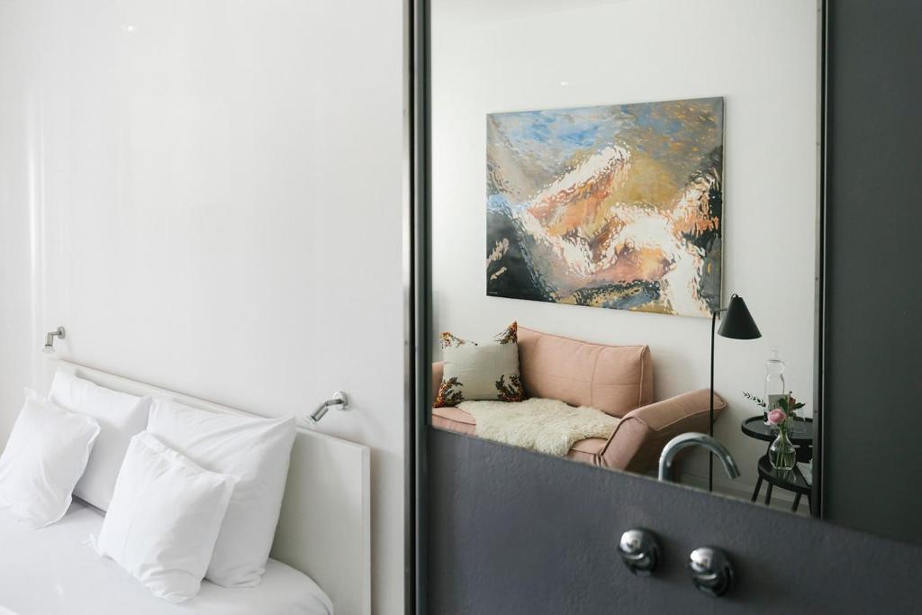 Mūsdienīga interjera attēls ar gaišu krēslu un mākslas darbu uz sienas, kas atspoguļojas spogulī. Telpa ir ērti iekārtota ar mīkstām spilveniem un dekoratīvu lampu.